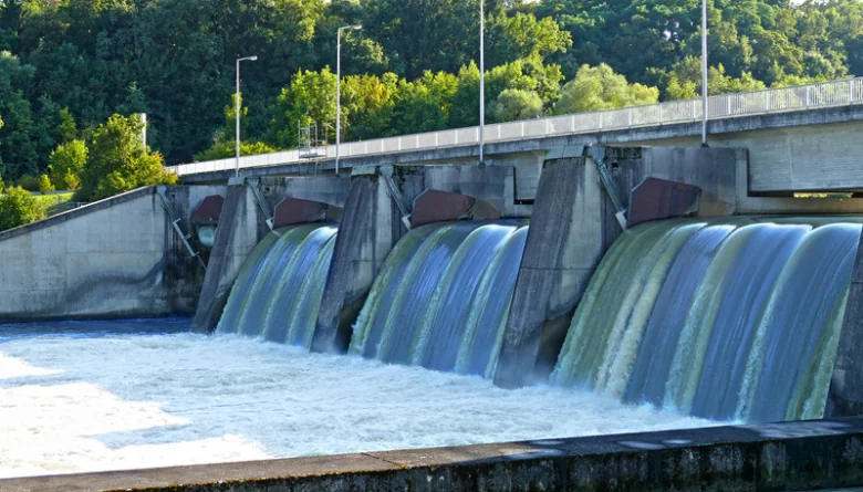 Water rushing through the spillway gates of a hydroelectric dam on a river lined with green trees