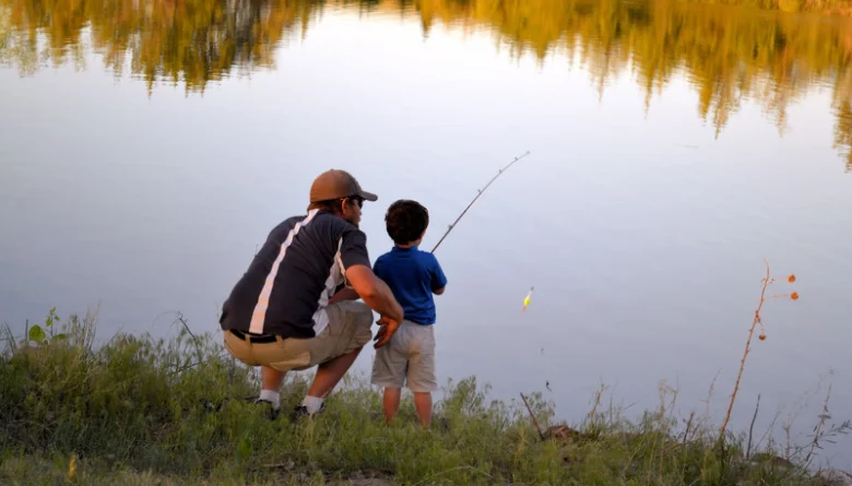 Father teaching his young son to fish from the grassy shore of a calm pond in summer